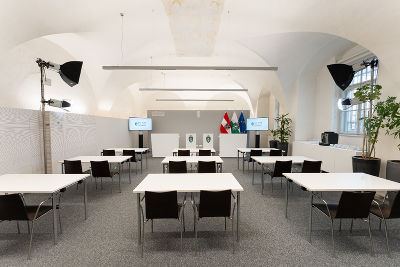Bildbeschreibung: Ein Raum für Pressekonferenzen mit Tischen und Stühlen, zwei Bildschirmen, einem Podium vor einer grauen Rückwand und drei Flaggen.