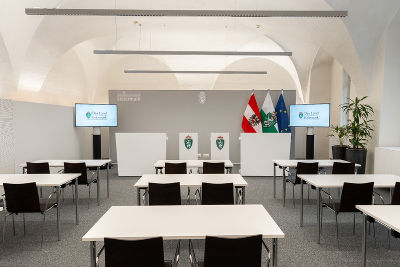 Bildbeschreibung: Ein Raum für Pressekonferenzen mit Tischen und Stühlen, zwei Bildschirmen, einem Podium vor einer grauen Rückwand und drei Flaggen.