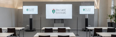 Bildbeschreibung: Ein Raum für Pressekonferenzen mit Tischen und Stühlen, einer grauen Rückwand, drei Bildschirmen und zwei freistehenden Mikrofonen vor dem Podium.